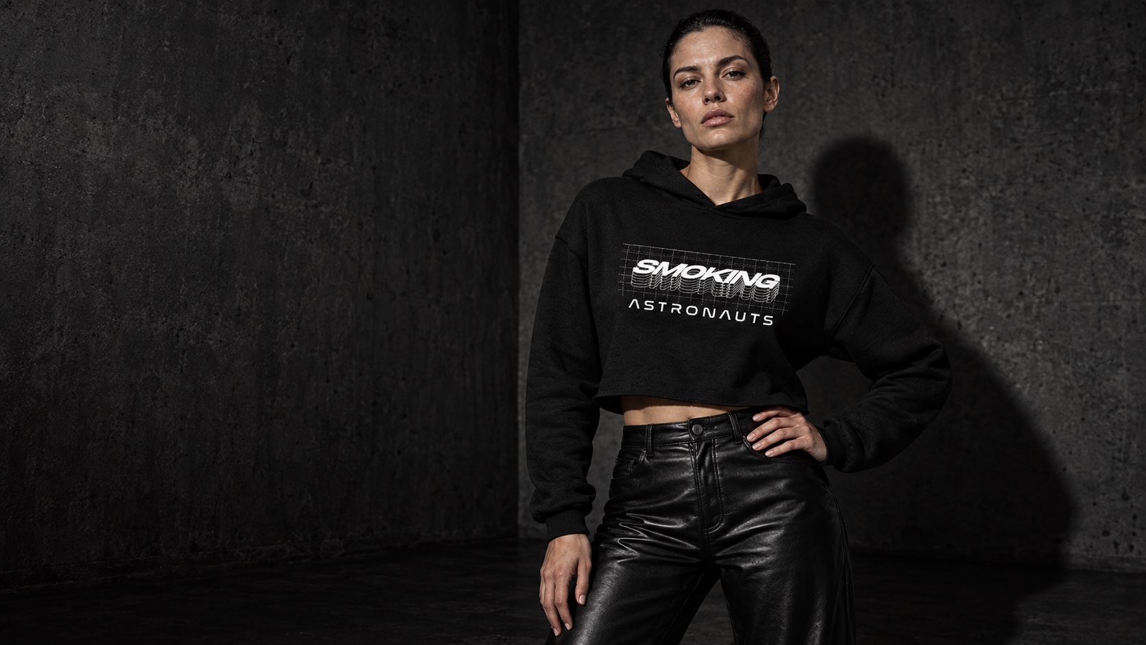 AI campaign concept: an adult model in the Cosmic Grid cropped hoodie, styled with black trousers against a charcoal wall